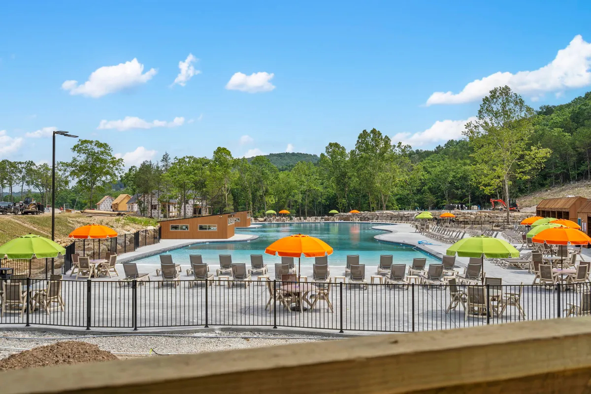 A lively outdoor scene featuring a swimming pool surrounded by colorful umbrellas, lounge chairs, and mature trees. The bright, cheerful mood invites families to enjoy swimming, sunbathing, and relaxing together under the sun.