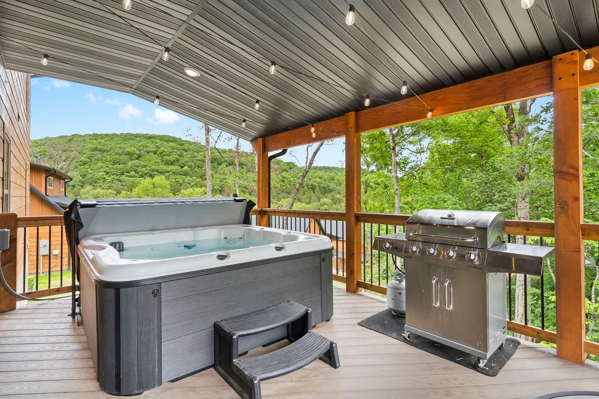 A wooden deck with a private hot tub overlooks scenic mountain views. The deck also features a grill and comfortable seating, perfect for relaxing or entertaining friends and family. The setting sun casts a golden glow, inviting guests to unwind in the warm bubbling water or enjoy the open-air atmosphere.