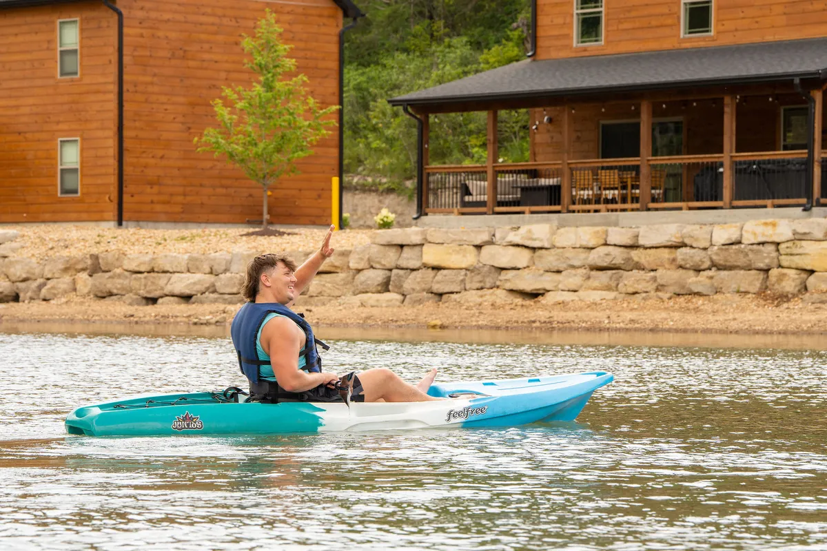 A playful lakeside image with a kayaker on the water, a nearby house, and lush greenery. The scene captures active family recreation such as kayaking, swimming, and relaxing by the lake.