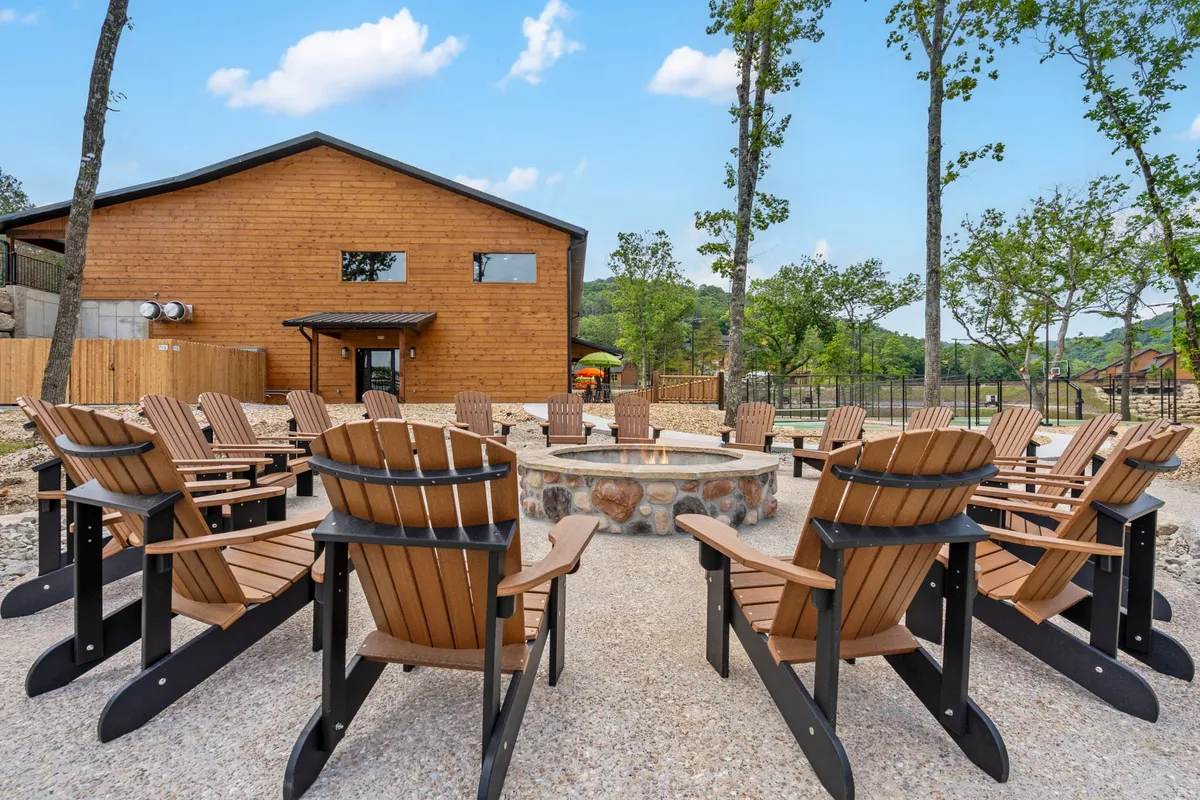 A fire pit surrounded by Adirondack chairs and nestled beside a wooden building with trees in the background. The scene suggests a classic campfire gathering spot, perfect for storytelling or unwinding after a day of activities.