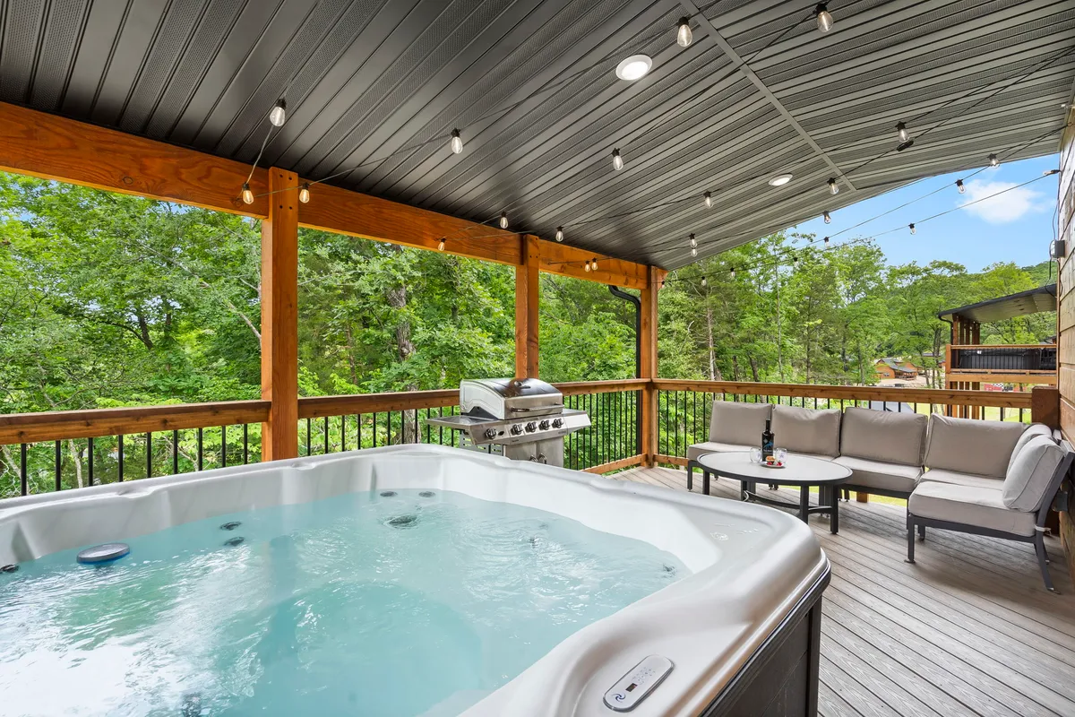 The image showcases the cabin’s spacious deck set against lush Ozark greenery, featuring a bubbling private hot tub, inviting outdoor seating, and a modern grill—all basked in the glow of a sunset. The tranquil setting highlights opportunities for relaxing soaks, lively cookouts, and peaceful mornings surrounded by nature.