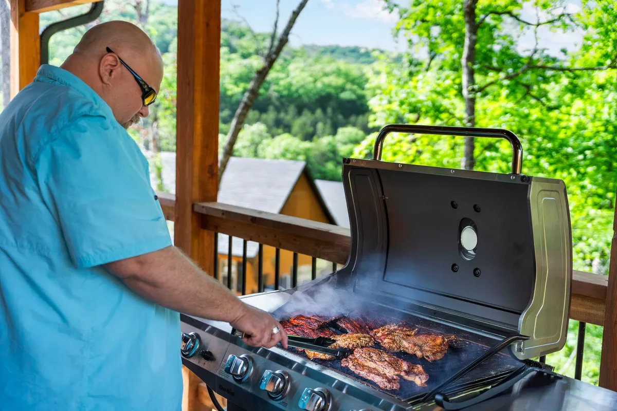 An inviting exterior scene with a man grilling outside, surrounded by lush greenery and cabins in the background. The atmosphere is casual and social, suggesting outdoor relaxation and memorable barbecues.