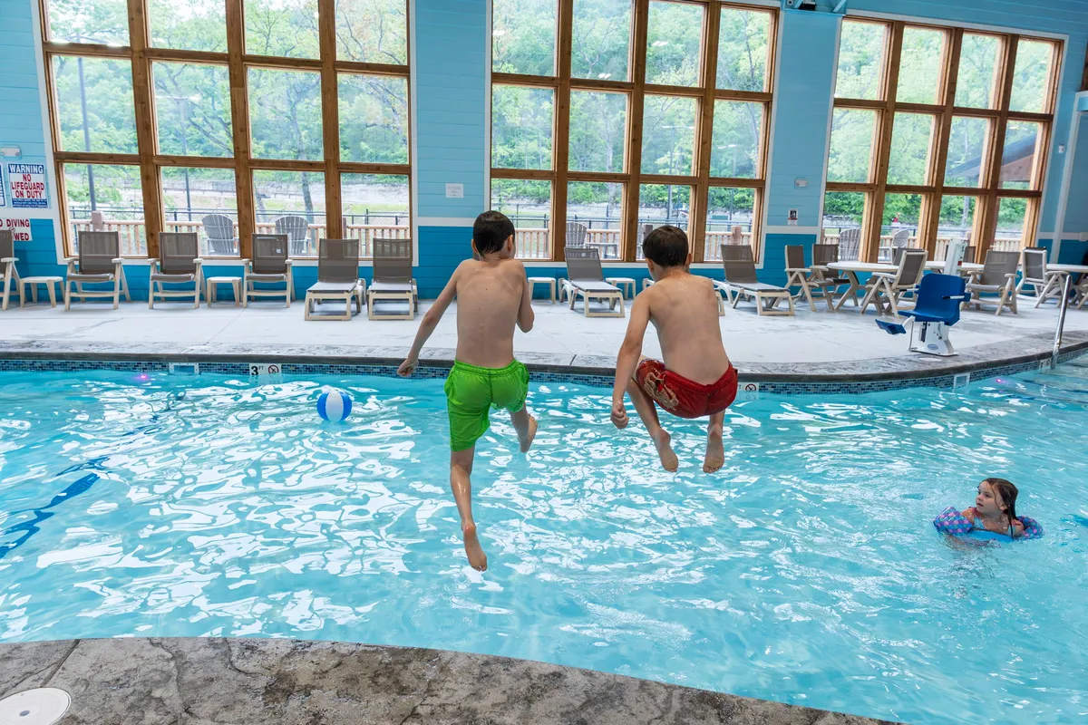 Children joyfully jumping into a sunlit indoor pool, with natural light streaming through large windows. The pool area is lively, filled with playful activity and the sound of laughter, evoking the sense of family fun and memories being made.