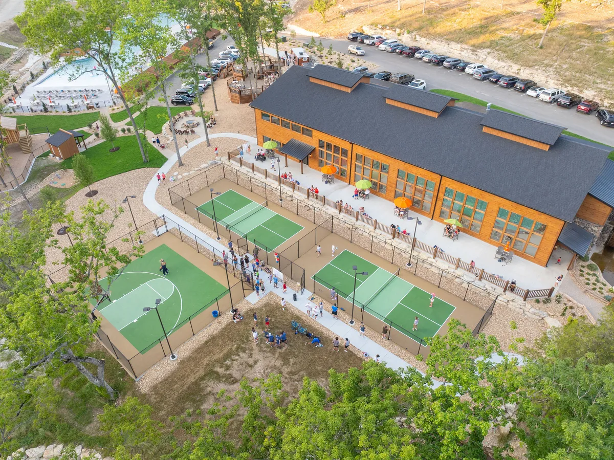 Spacious outdoor sports courts adjacent to the main building and gathering areas, framed by greenery and a vibrant, communal atmosphere. Groups of people are seen enjoying basketball and tennis, emphasizing activity and togetherness.