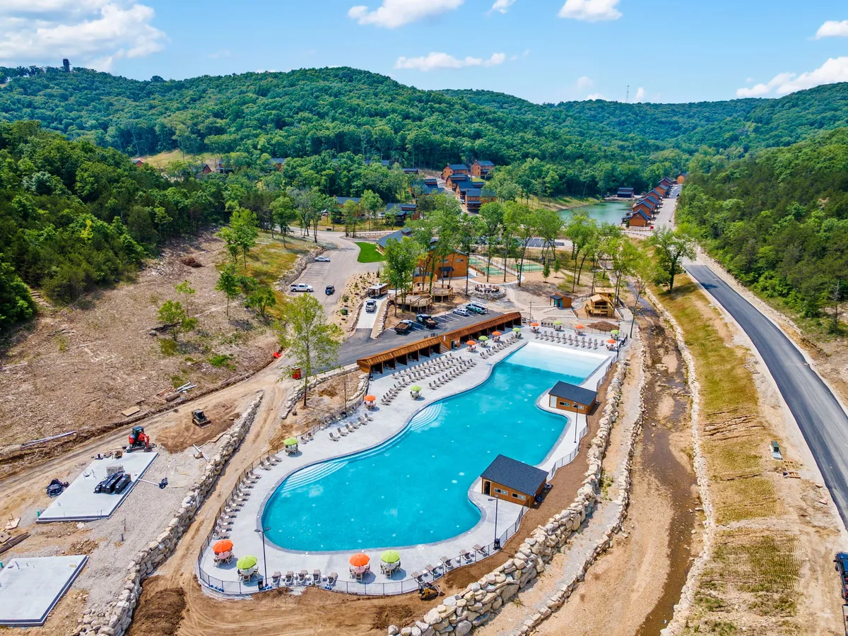 A large, inviting outdoor pool surrounded by lounge chairs and a wooden structure with seating, set against a backdrop of lush green hills. The setting is relaxed and cheerful, suggesting summer gatherings and outdoor fun.