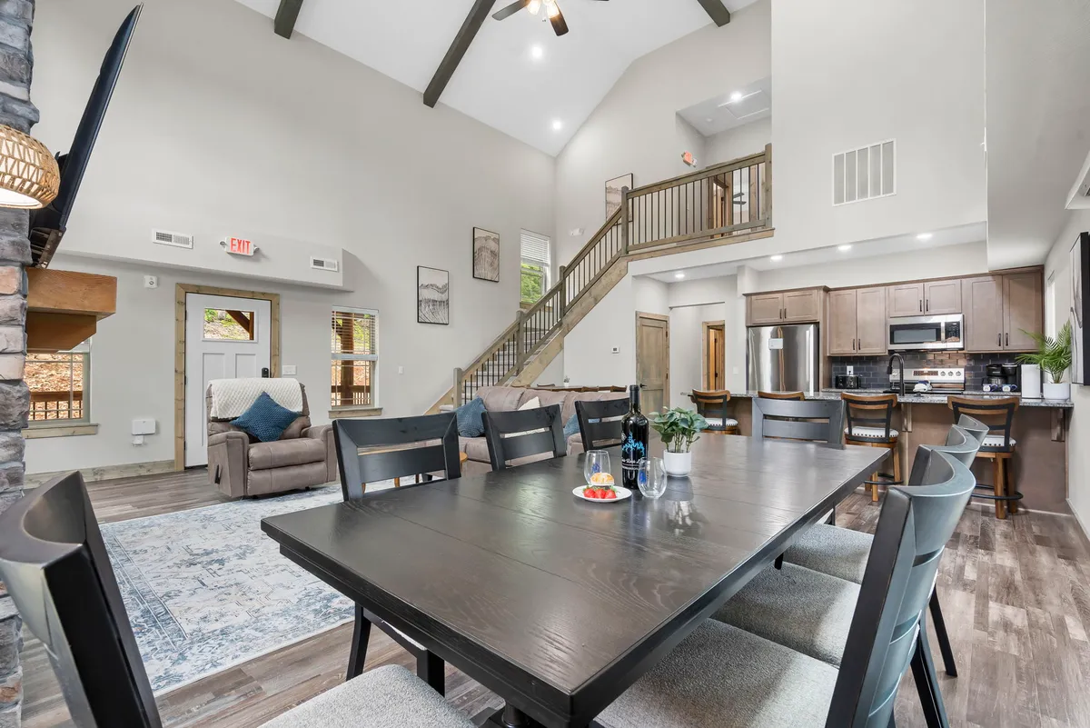 This open-plan interior highlights a spacious dining table set amid a kitchen area and living space with a sofa, with a staircase and large windows providing a sense of light and openness. The arrangement invites family gatherings, shared meals, and relaxed entertaining in a harmonious flow.